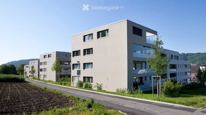 Modern multi-story residential building with large windows and balconies, surrounded by greenery and a paved road.