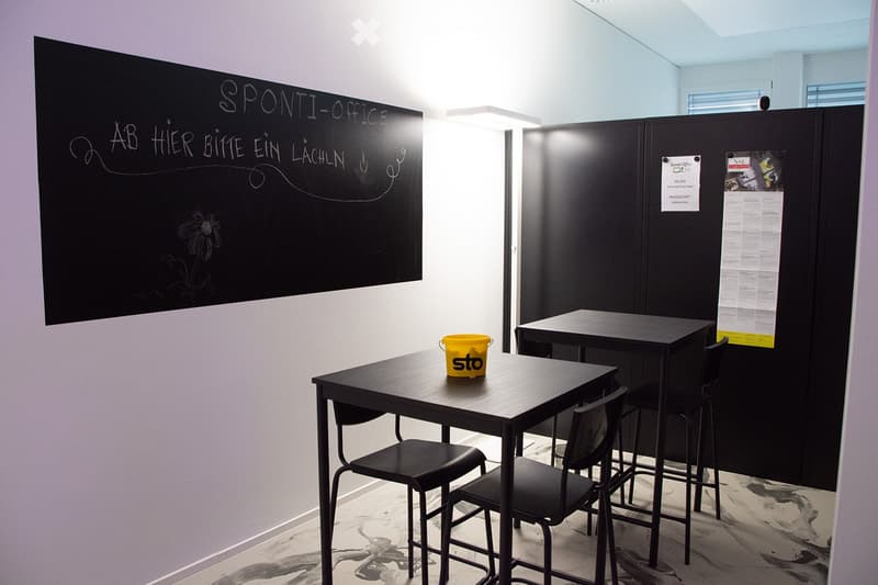 Office space with two black tables, chairs, a yellow stool, a blackboard with text, and posters on the wall.