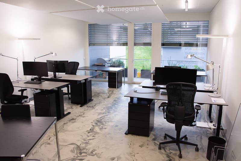 Modern office space with multiple desks, ergonomic chairs, computers, keyboards, and lamps. Large windows with blinds provide natural light.