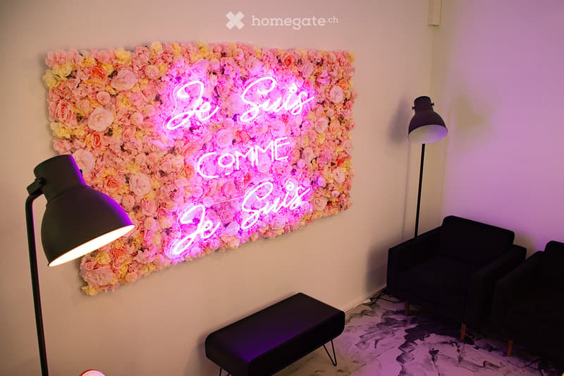 A stylish living room with a floral wall display, neon sign reading "Je Suis Come Je Suis", a black sofa, a small black stool, and two floor lamps.