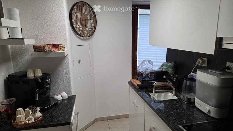 Modern kitchen with granite countertops, white cabinets, and various appliances including a coffee maker, microwave, and oven. Shelves hold cups and a decorative clock is mounted on the wall.