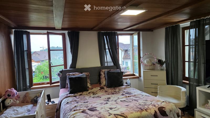 Cozy bedroom with a large bed, colorful pillows, nightstand, and a view of the neighborhood through large windows.