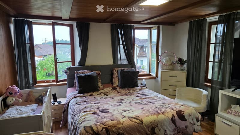 Cozy bedroom with a double bed, nightstand, chair, and a basket on a dresser. Large windows offer a view of the neighborhood.