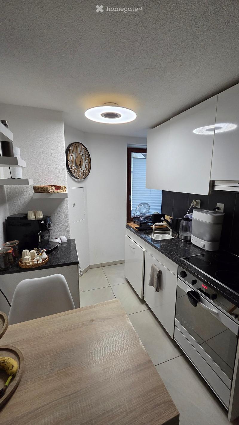 Modern kitchen with white cabinets, black countertops, and stainless steel appliances. Features a wooden dining table, a chair, and a clock on the wall.