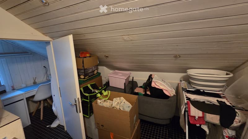 A small, cluttered room with a desk, chair, storage boxes, a bathtub, and a clothing rack. The space appears to be used for storage and possibly as a workspace.