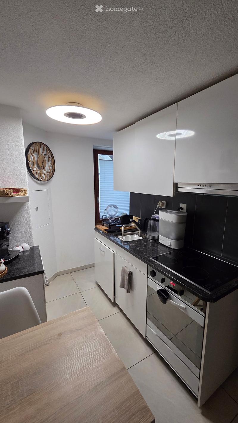 Modern kitchen with white cabinets, black countertops, and stainless steel appliances including an oven, stove, and sink. A wooden dining table is visible in the foreground.