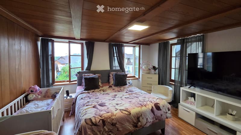 Cozy bedroom with wooden ceiling, large windows, a double bed with floral bedding, a TV stand, a small chair, and a crib with toys.