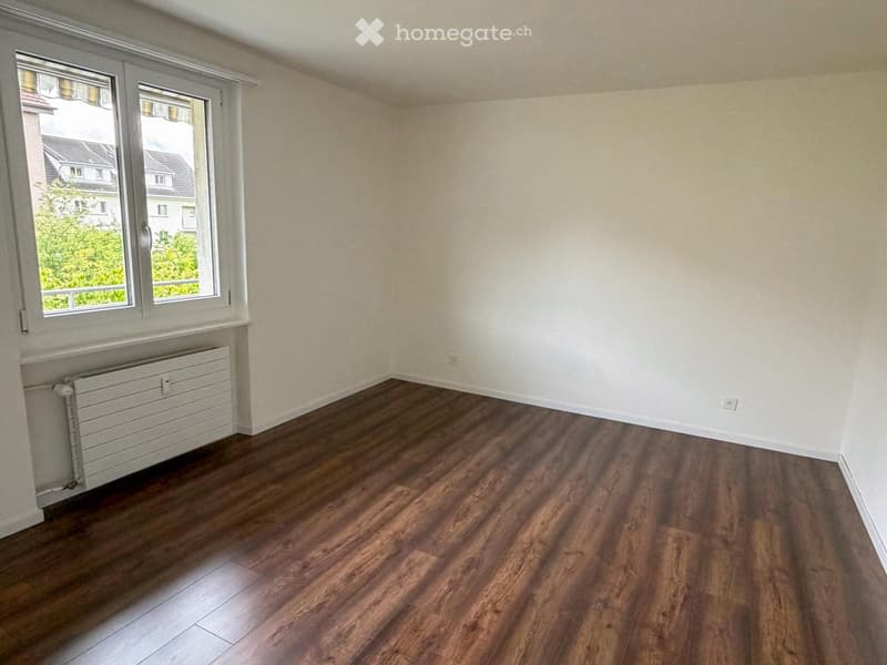 Empty room with wooden flooring, white walls, and a window with a view of neighboring houses and greenery.