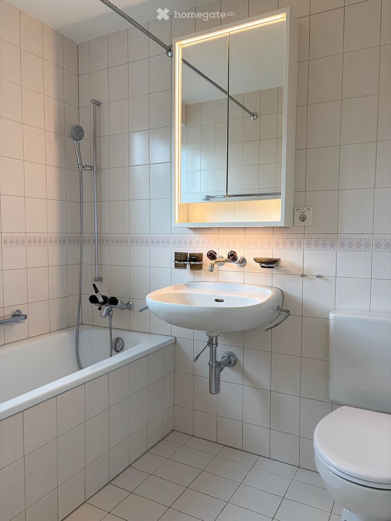 Modern bathroom with a bathtub, sink, toilet, and a large mirror with built-in lighting. The walls are tiled in a light beige color, and the floor is tiled in a light gray color.