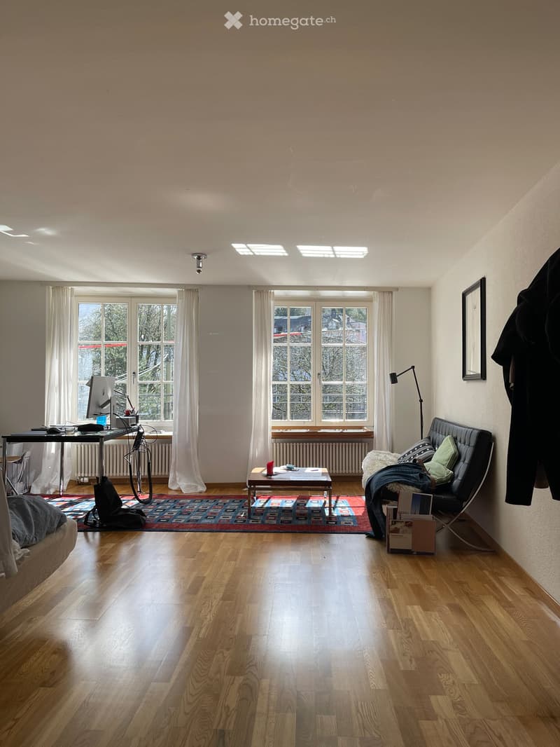 Bright, open living space with wooden flooring, large windows, a sofa, coffee table, desk with laptop, and a floor lamp.
