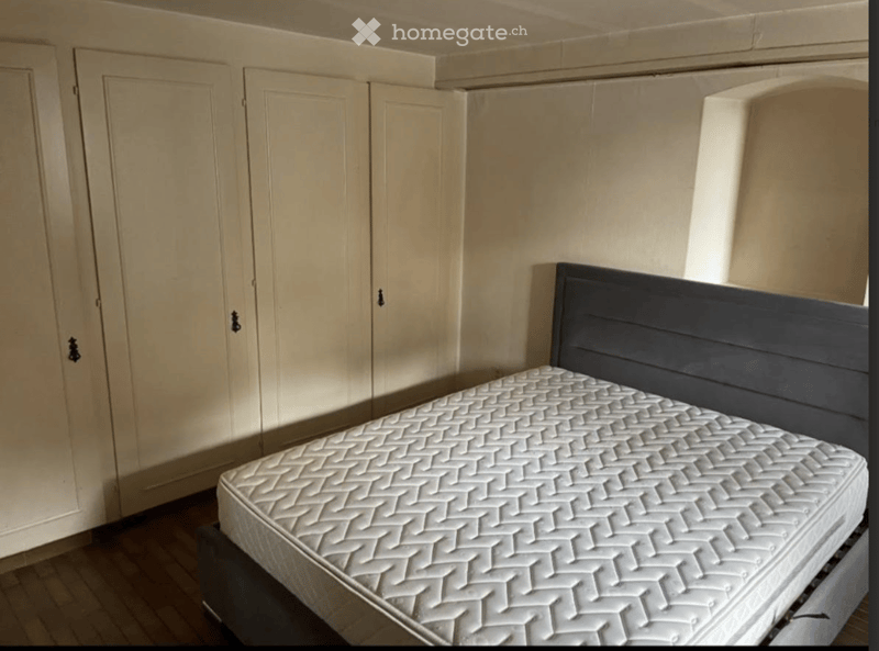 Bedroom with a single bed and a wardrobe. The bed has a dark headboard and a white quilted mattress. The wardrobe has double doors and is light-colored.