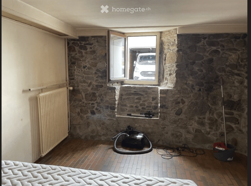 A small, rustic room with exposed stone walls, a tiled floor, and a single bed. A window with an open shutter reveals a parked vehicle outside. Cleaning supplies and a vacuum cleaner are visible on the floor.