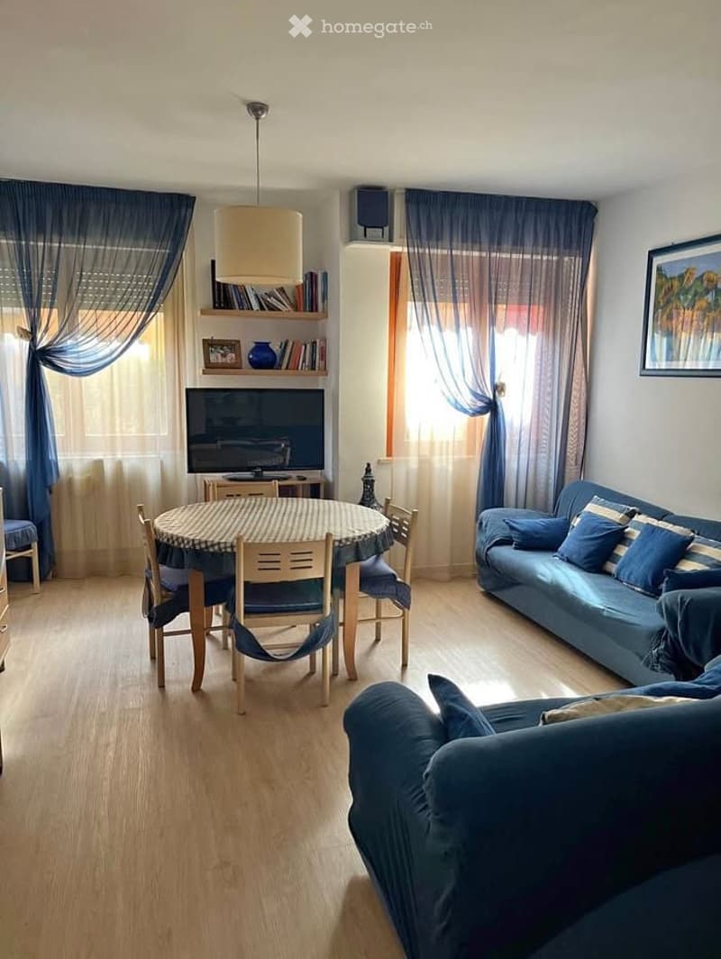 A modern living and dining area with a blue sofa, a round dining table with chairs, a television, a bookshelf, and large windows with curtains.