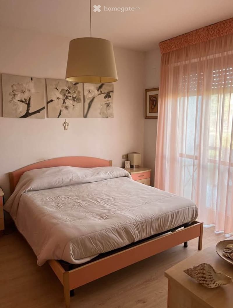 Cozy bedroom with a double bed, nightstand, lamp, and artwork on the wall. Light-colored curtains cover the window, and a small table with a decorative item sits near the bed.