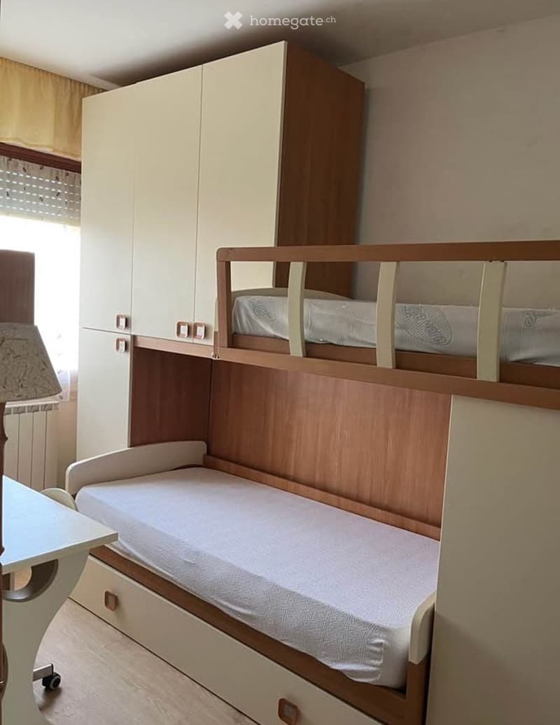A compact bedroom with a bunk bed, a wardrobe, a small table, and a lamp. The room features light-colored furniture and a window with curtains.