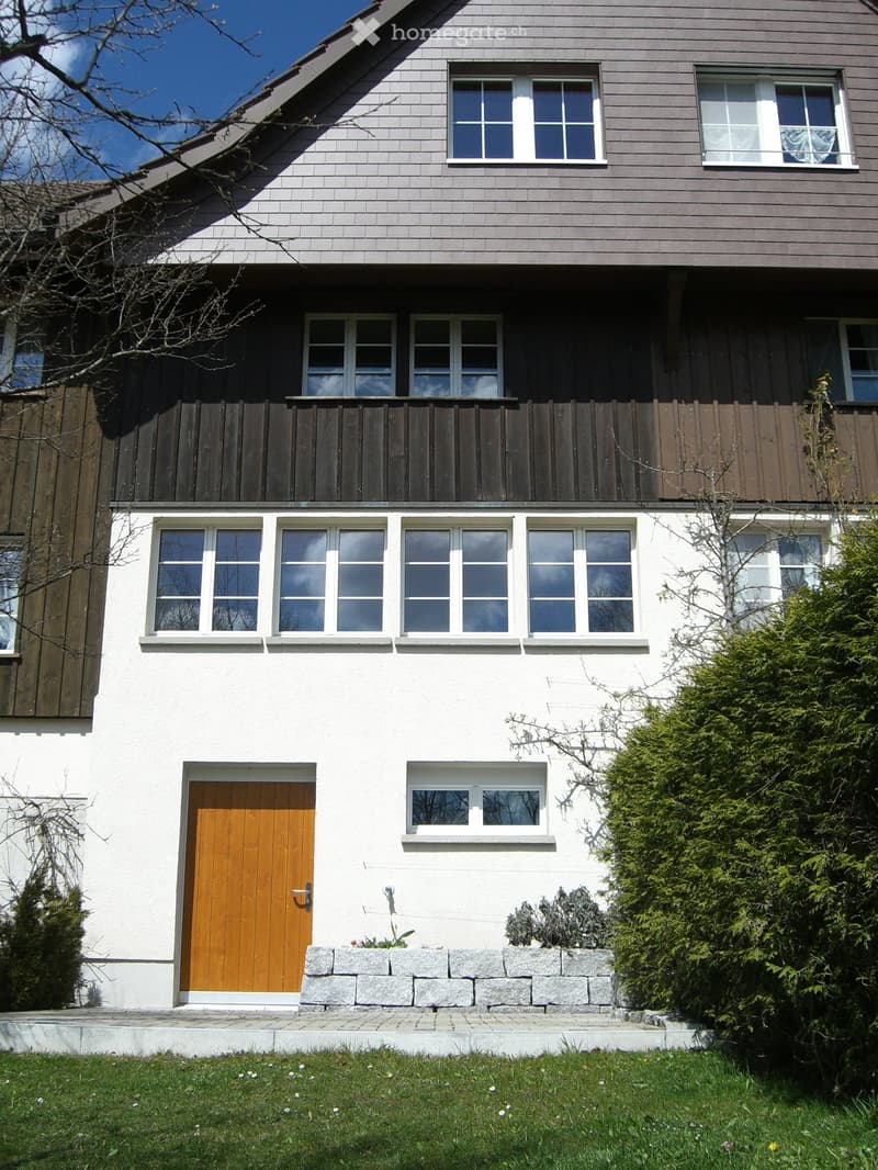 Two-story house with white walls, multiple windows, wooden front door, and a small garden area with stone steps.