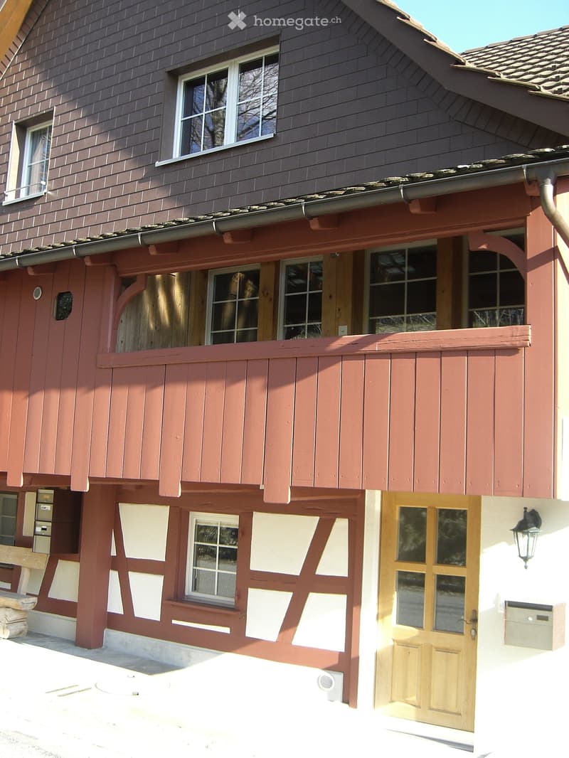 Two-story house with red wooden siding, white-framed windows, and a wooden front door. Features a covered entrance area with wooden beams and a small bench on the left side.