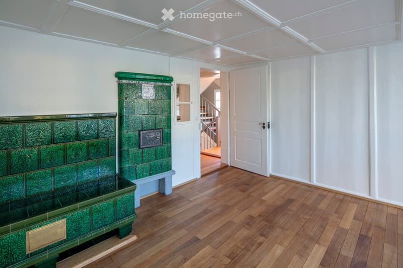 A well-lit hallway with wooden flooring, white walls, and a decorative green fireplace on the left side. A white door is visible on the right, leading to another area.