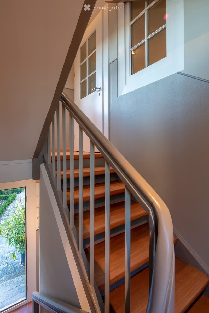 Modern staircase with wooden steps and a metal railing, leading to an upper level. The walls are painted in neutral tones, and there is a window at the bottom of the stairs offering a view of the garden.