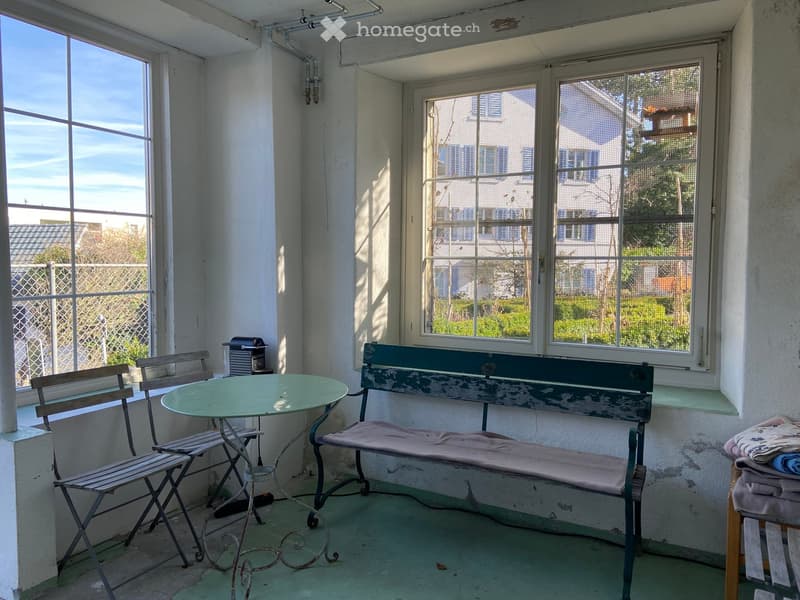 A furnished balcony with a round table, two chairs, a bench, and a small appliance, overlooking a residential area.