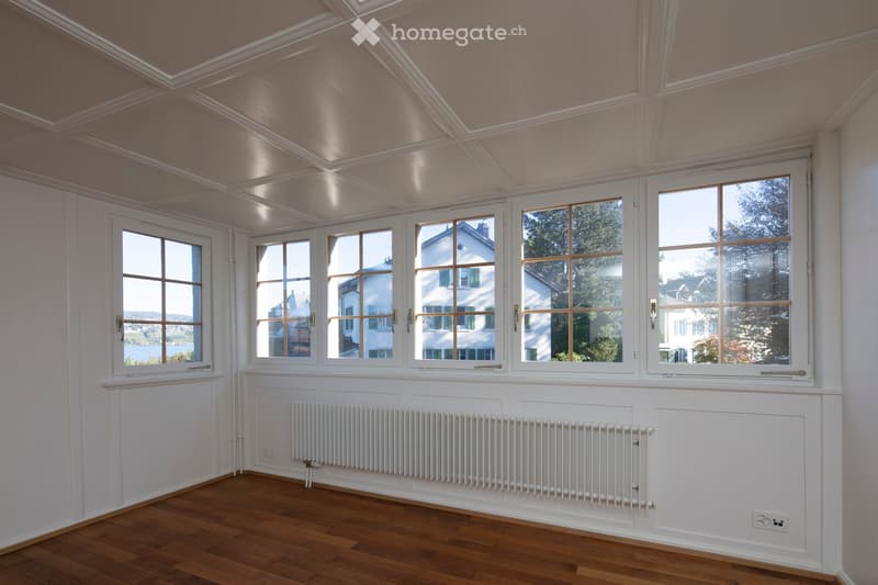 Bright, empty room with wooden flooring, white walls, large windows, and a white radiator. The ceiling features decorative molding and skylights.