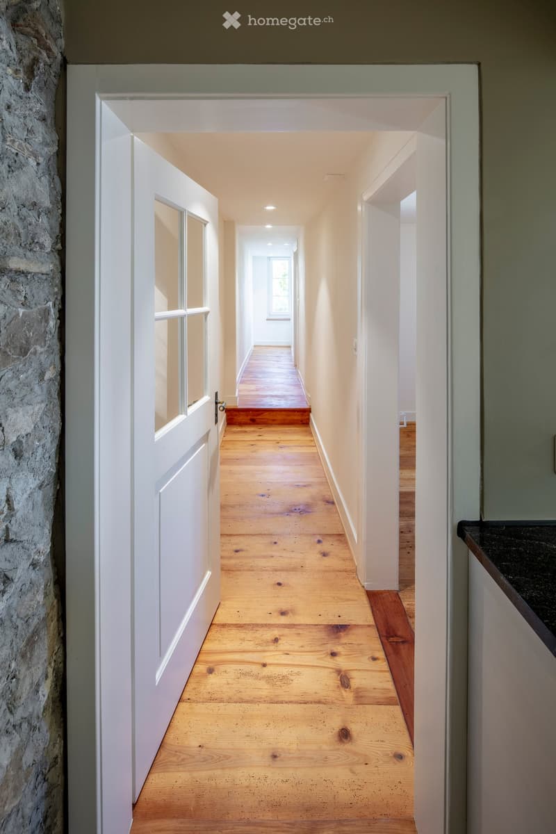A long, narrow hallway with wooden flooring, white walls, and a stone wall on one side. The hallway has recessed lighting and a white door at the end.