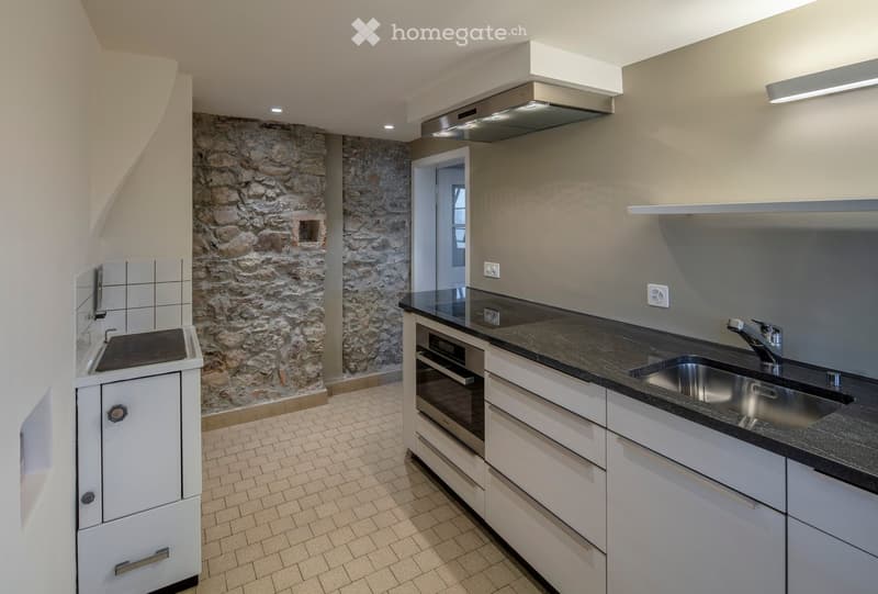 Modern kitchen with white cabinets, black countertop, stainless steel sink and faucet, built-in oven, and a range hood. Exposed stone wall adds texture.