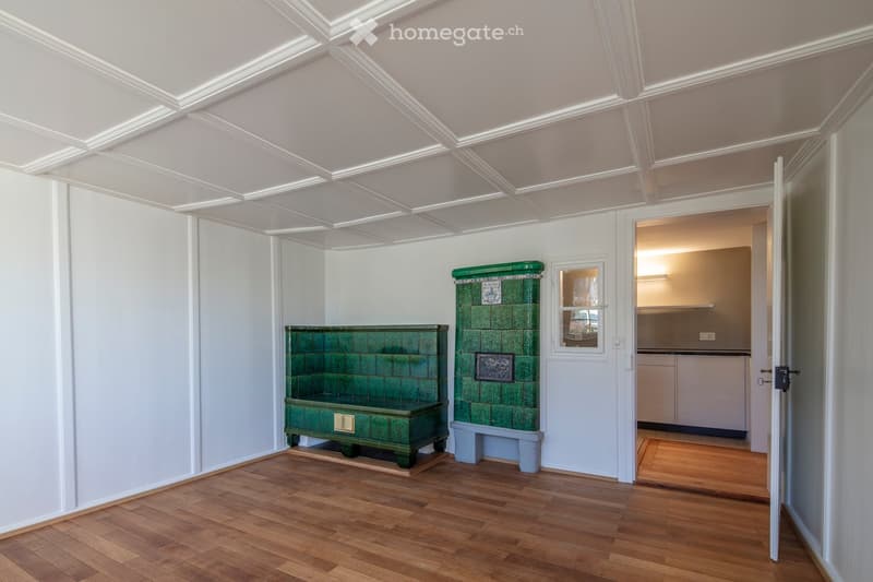 A room with wooden flooring, white walls, and a decorative green bench and fireplace. An adjacent kitchen area is visible through an open doorway.
