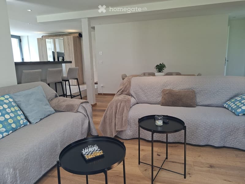 Modern living room with a sectional sofa, two black side tables, a chessboard, and a small kitchen area with bar stools.