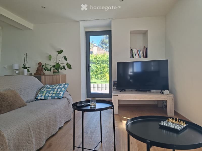 Modern living room with a grey sofa, coffee table, TV, and a chessboard on a round table. Features large windows with a view of greenery outside.