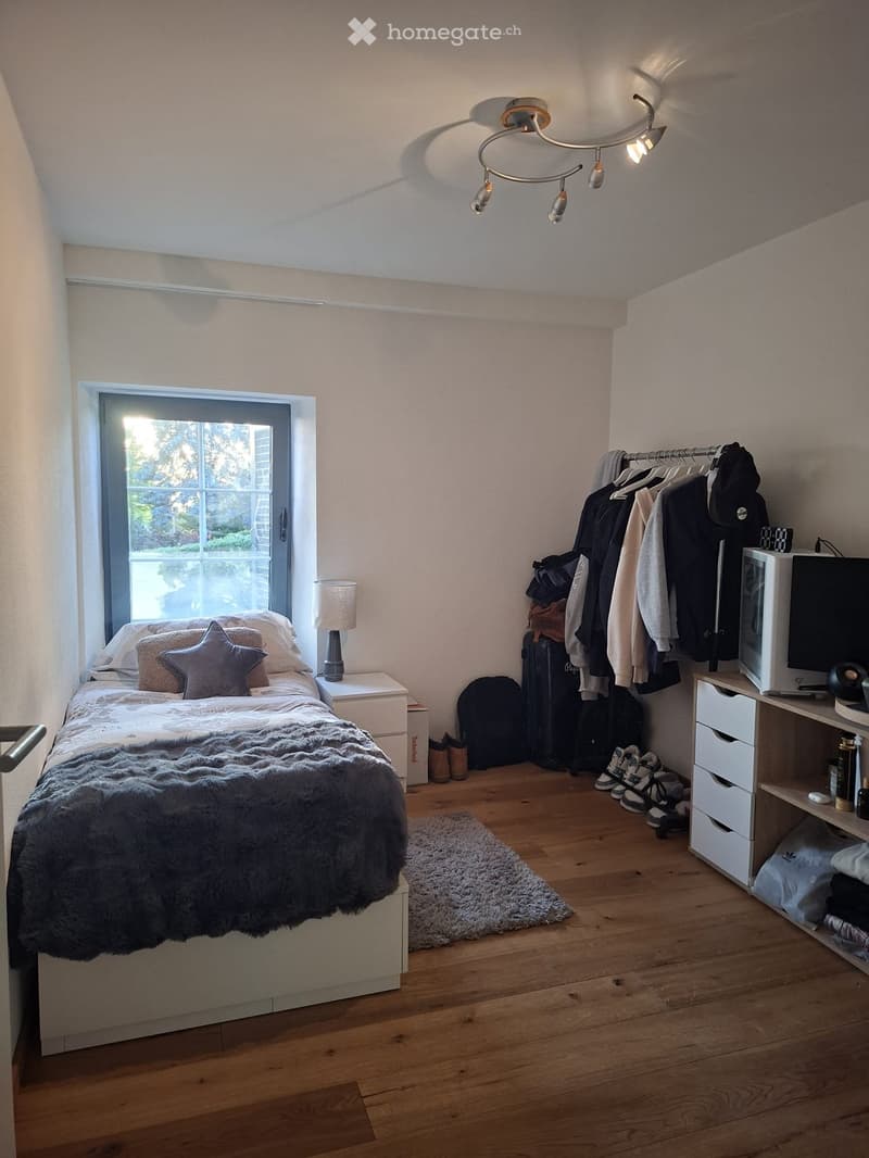 Cozy bedroom with wooden flooring, a bed with a fur throw and star-shaped pillows, a nightstand with a lamp, a clothing rack with jackets and bags, a drawer unit with a screen, and a small rug near the bed.
