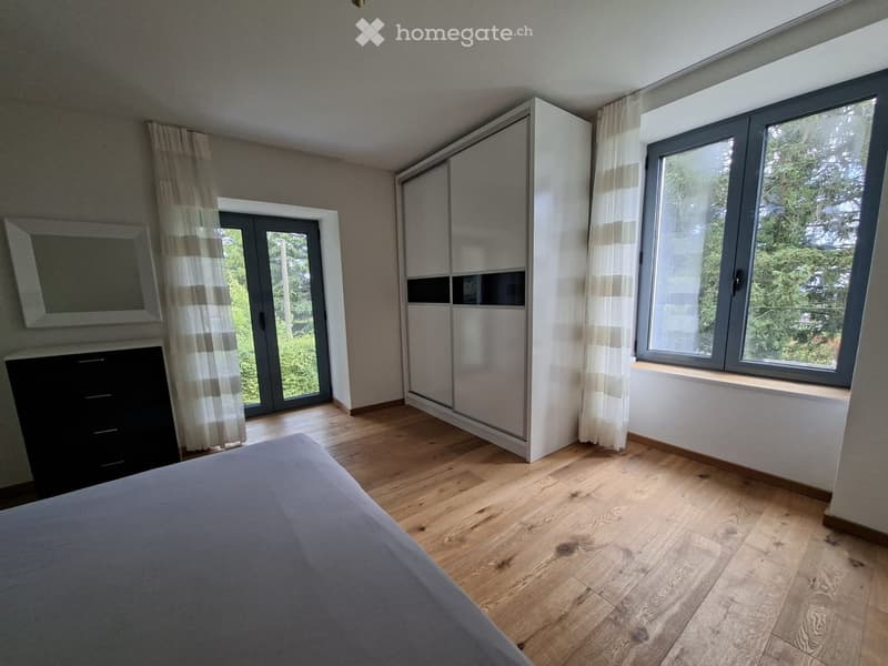Modern bedroom with wooden flooring, large windows, sliding doors, and a bed with grey bedding.