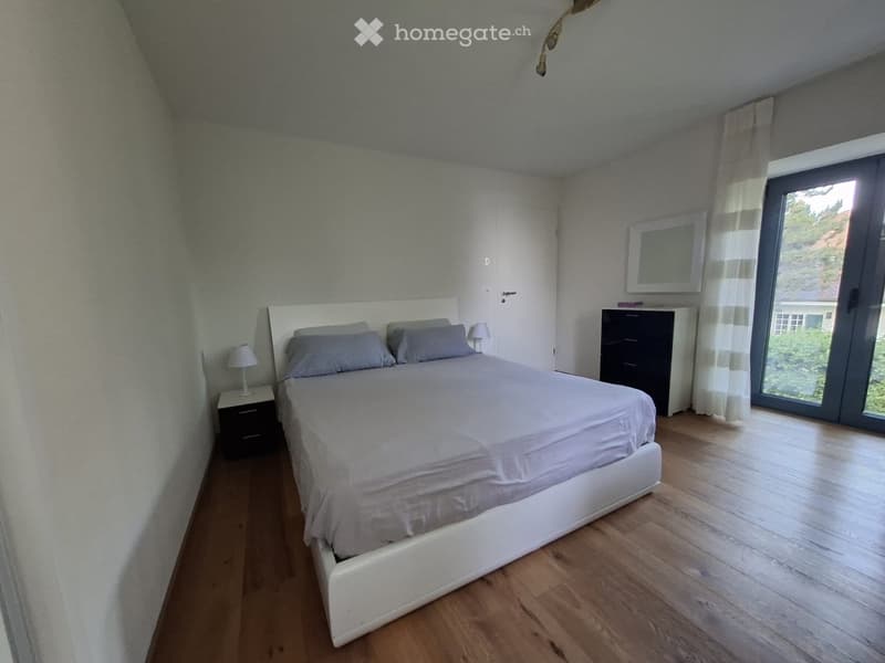 Modern bedroom with a white bed, grey bedding, wooden floor, nightstand with lamp, and a large window with curtains.