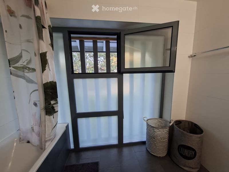 Bathroom with a shower curtain, a laundry basket, and a trash bin near a window with frosted glass panels.