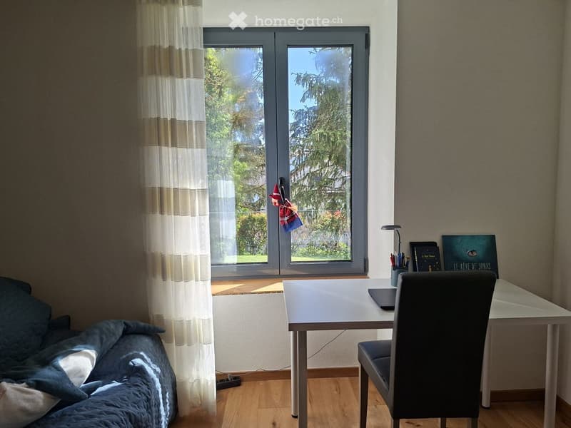 A cozy, well-lit room with a sofa, a desk with a laptop, and a chair. The room features large windows with curtains, a view of greenery, and a small decorative item hanging on the window.