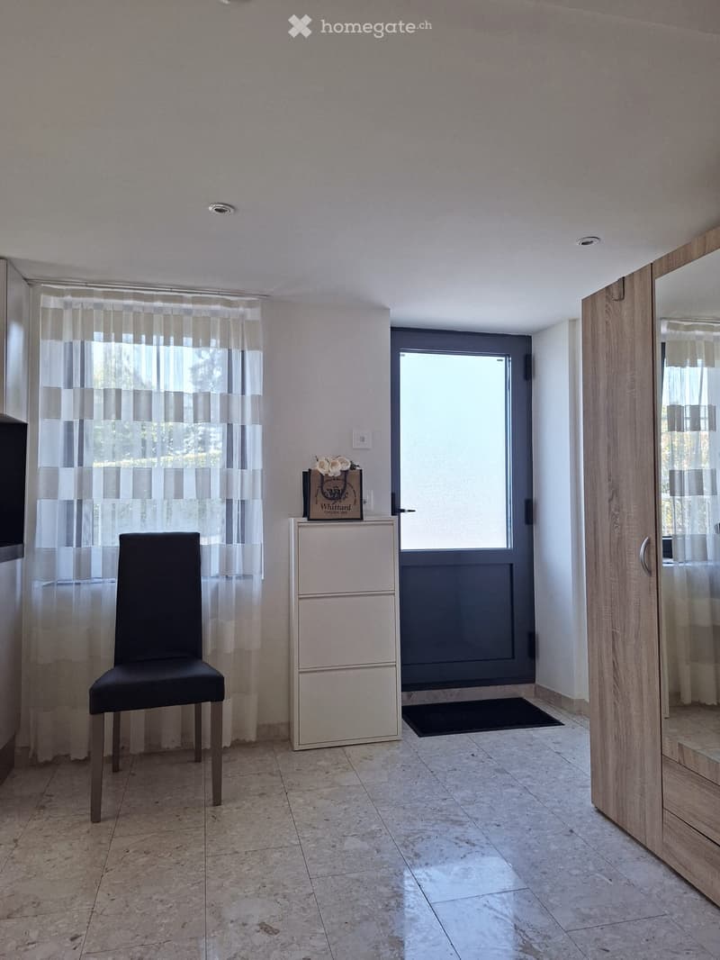 A modern hallway with a tiled floor, a black chair, a small cabinet with a decorative box on top, and a large mirror on the right side.