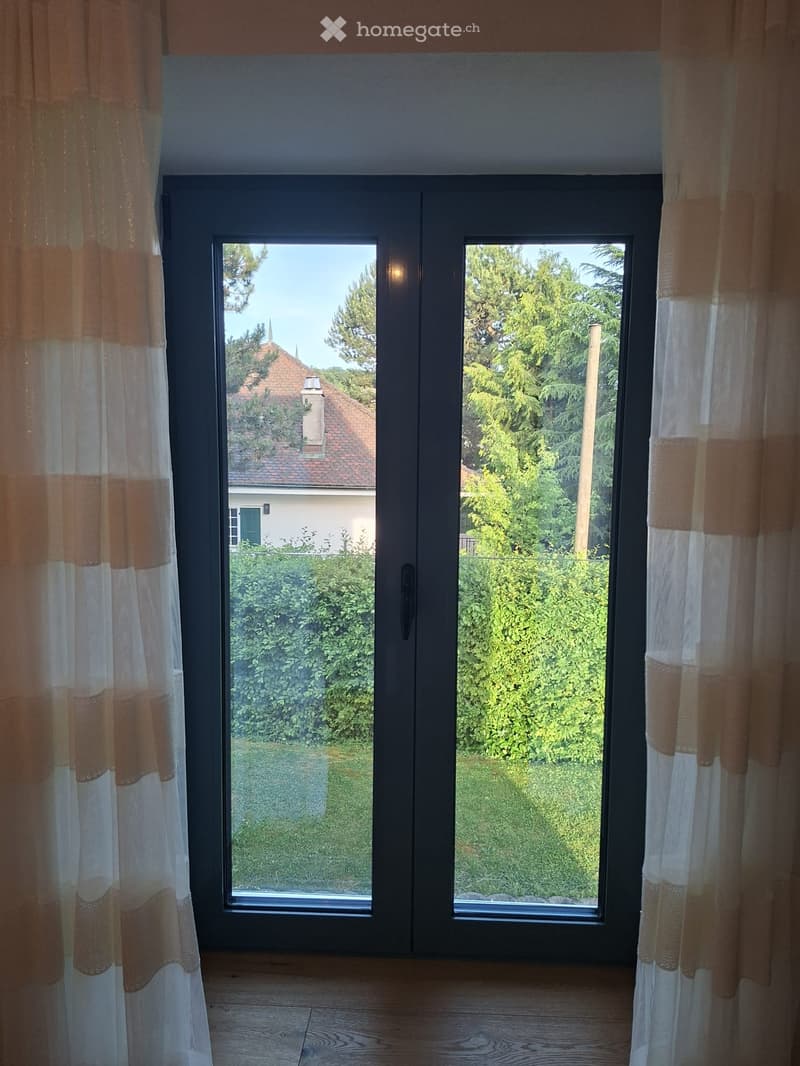 Empty room with wooden flooring, large double doors leading to a garden, and sheer curtains on either side.