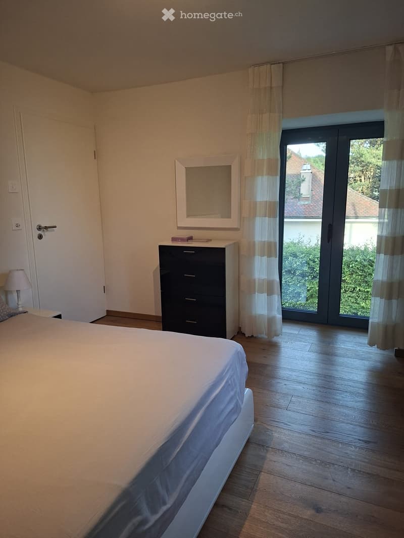 Modern bedroom with a large bed, nightstand with lamp, mirror, and a drawer unit. Large sliding glass door with curtains leads to a view of greenery.