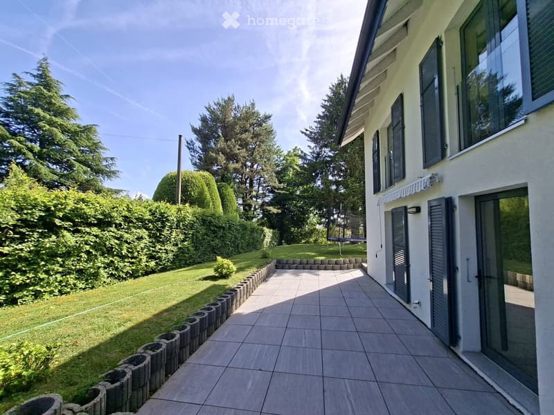 Garden area with tiled patio, neatly trimmed hedges, and a small grassy lawn, adjacent to a modern house with large windows and shutters.