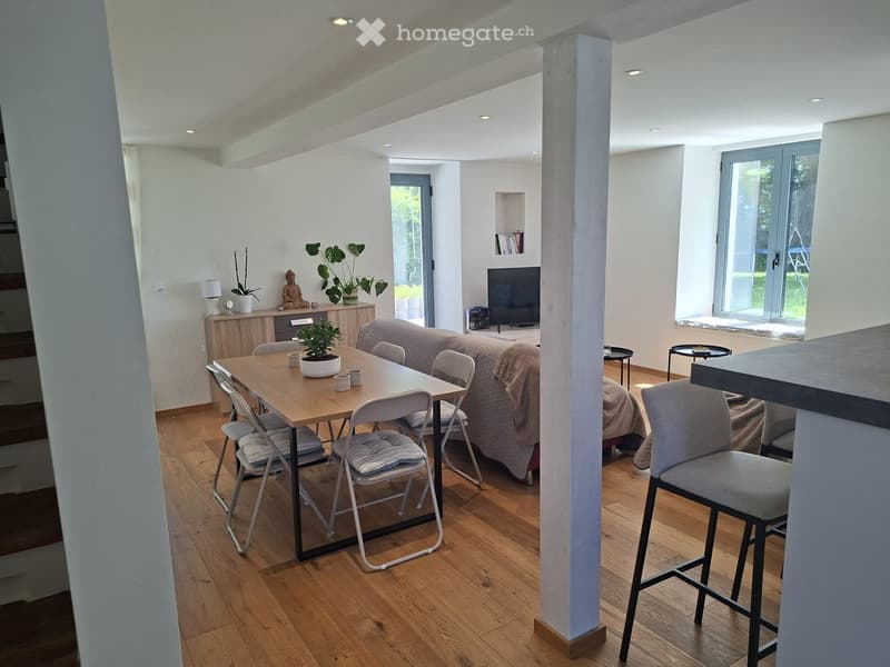 Open-plan living and dining area with wooden flooring, a sofa, dining table with chairs, a television, and decorative plants.