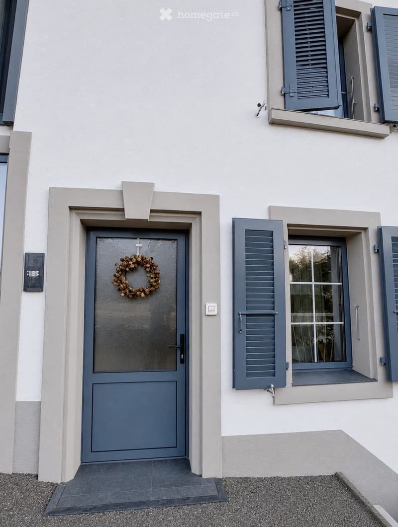 Front entrance of a house with a grey door adorned with a decorative wreath, flanked by windows with grey shutters.
