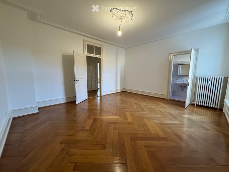 Empty room with wooden herringbone flooring, white walls, and a decorative ceiling light. Two open doors lead to adjacent spaces, one showing a bathroom with a sink and radiator.