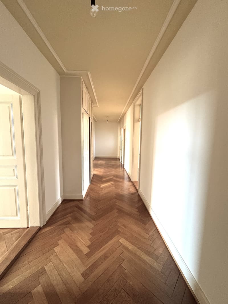 A long, narrow hallway with herringbone-patterned wooden flooring, white walls, and several doors on both sides.