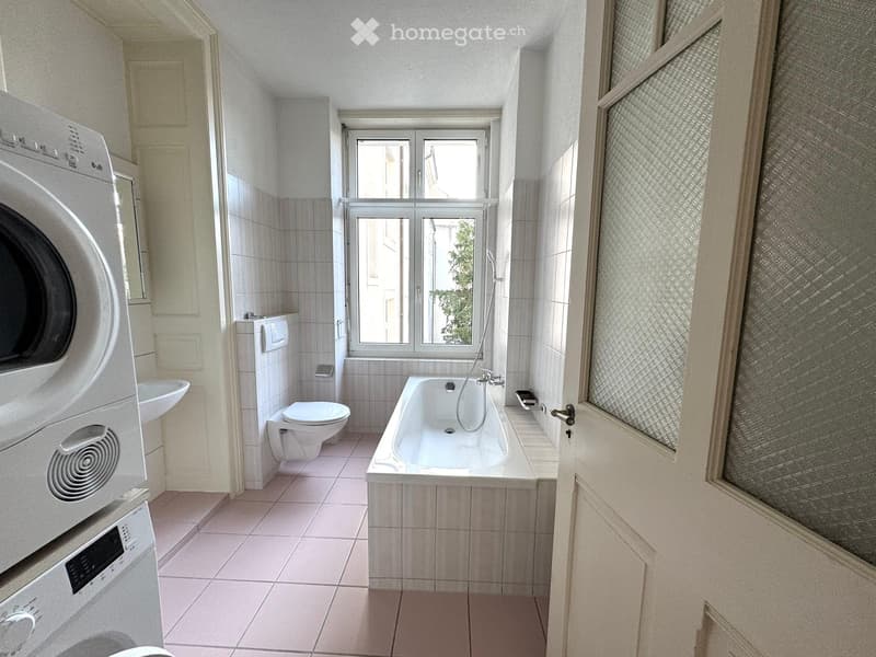 Bathroom with a bathtub, toilet, sink, and washing machine. The room has tiled walls and floor, with a window providing natural light.
