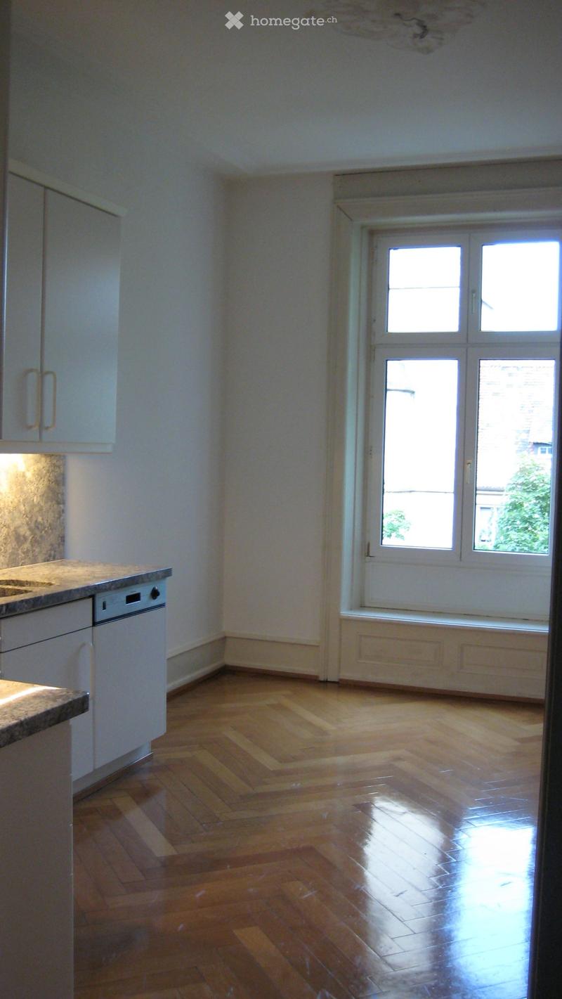 Empty kitchen with wooden flooring, white cabinets, a sink, and a dishwasher. Large windows allow natural light to enter.