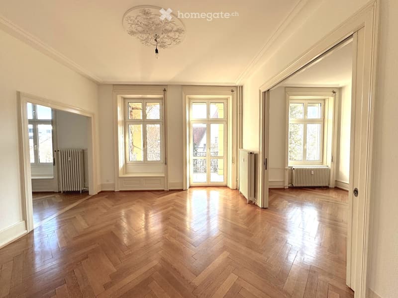 Empty room with wooden herringbone flooring, white walls, large windows, radiators, and an ornate ceiling light fixture.
