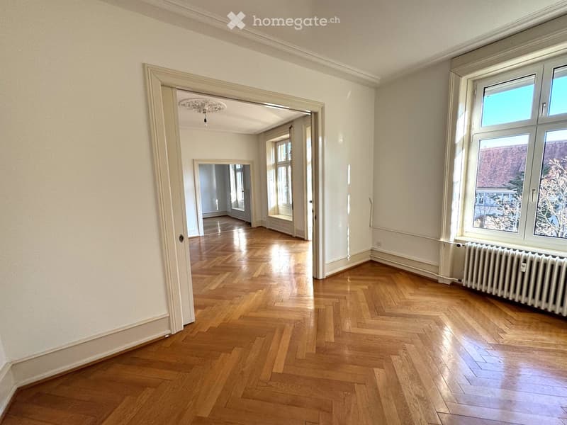 Empty room with wooden herringbone flooring, white walls, large windows, and a radiator. The room has an open doorway leading to another room with similar flooring and white walls.