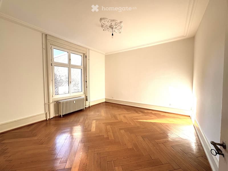 Empty room with wooden herringbone flooring, white walls, large window, radiator, and ornate ceiling light fixture.