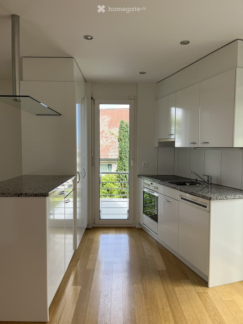Modern kitchen with light-colored cabinets, granite countertops, wooden flooring, and a view of a balcony and garden through a glass door.