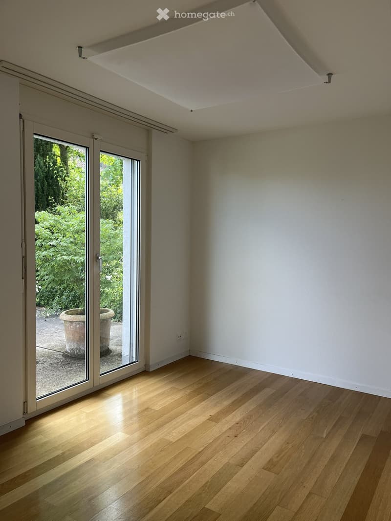 Empty room with light wooden flooring, white walls, and large sliding glass doors leading to a garden area.